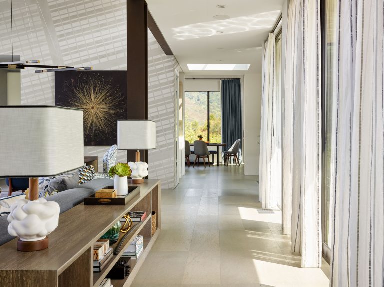 Bright, modern living space with floor-to-ceiling windows draped with sheer curtains, letting in natural light. Neutral décor, a gray sofa, decorative lamps, and a shelf fill the foreground, with art on the wall and a distant seating area visible.