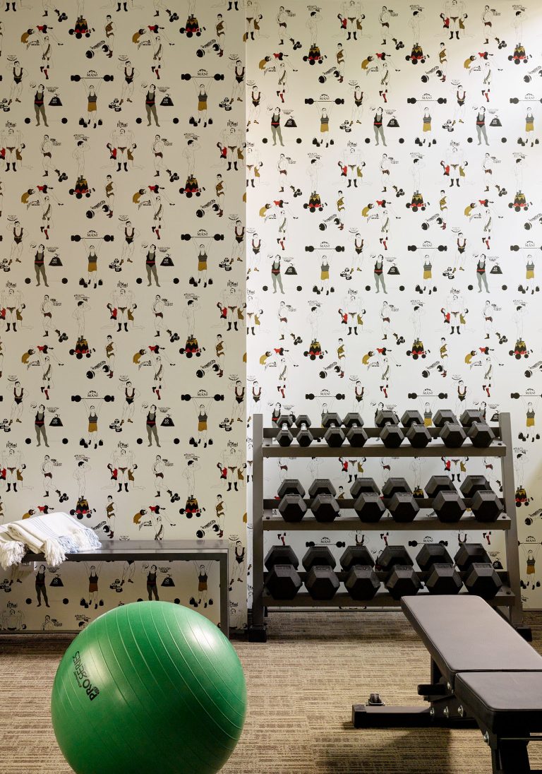 A gym corner with a green exercise ball, a weight bench, a rack of dumbbells, and a white towel on a metal bench. The walls feature whimsical wallpaper with cartoon animals lifting weights and exercising, creating a playful atmosphere.