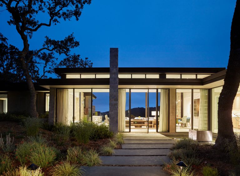 Modern, single-story house at dusk with large glass windows and doors, glowing warmly from interior lights. A stone pathway leads to the entrance, framed by minimalist landscaping and trees. The flat roofline and open living spaces are visible through the glass.