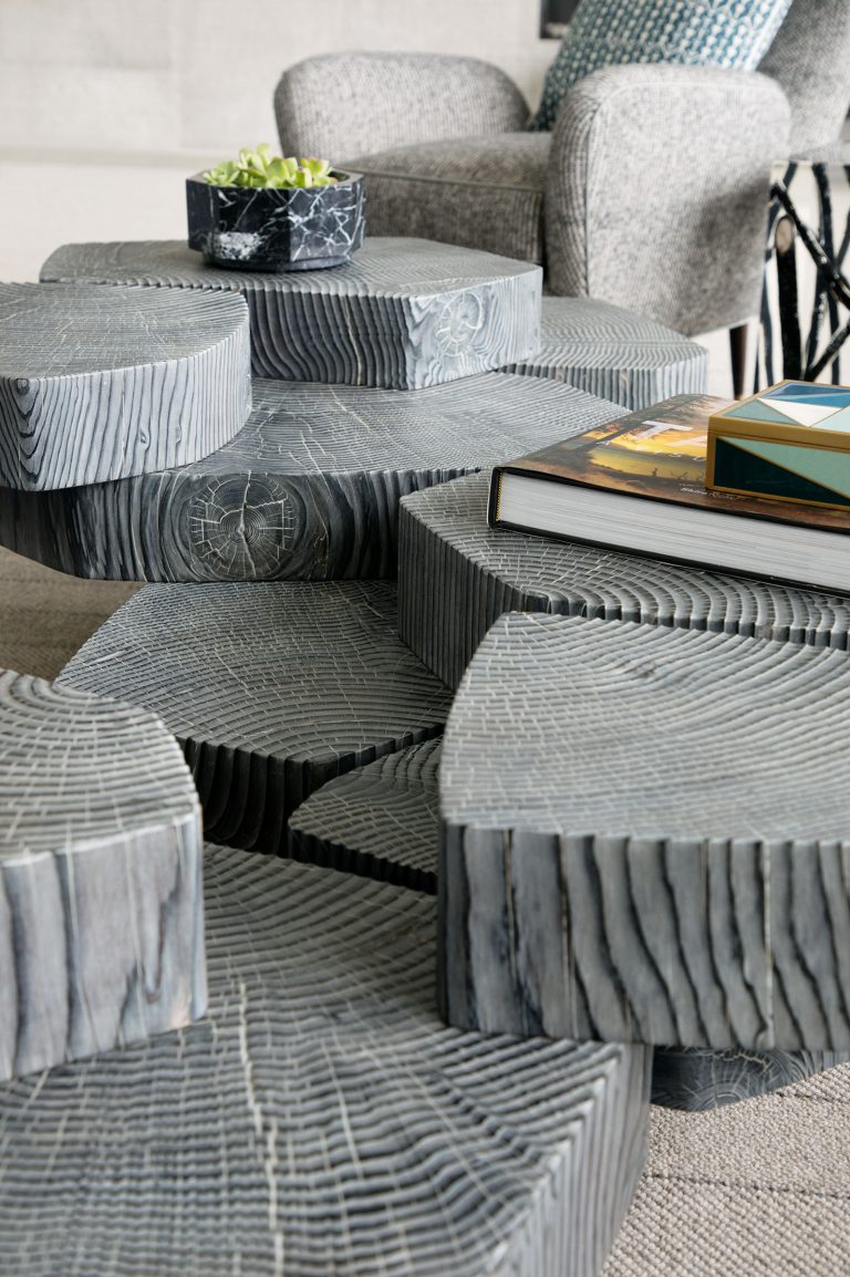 Close-up of a modern coffee table made from several large, round, dark gray wooden segments with visible tree rings and texture. On top are a small plant in a marble pot and a couple of hardcover books. A textured gray armchair is in the background.