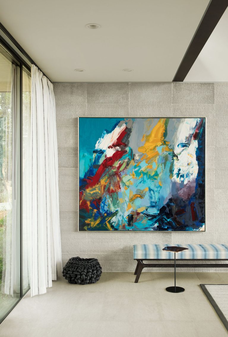 A large abstract painting with blue, white, red, orange, and yellow brushstrokes hangs on a gray stone wall above a striped blue bench and a small black round table. White curtains cover a glass door on the left; a black woven pillow sits on the floor.