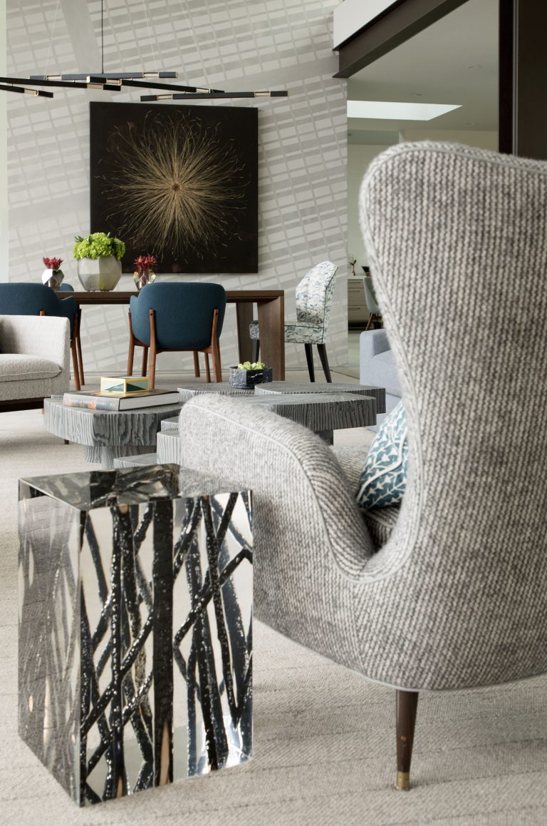 Modern living room with textured gray armchairs, a mirrored side table with tree-branch patterns, round coffee tables, and a dining area in the background. Large abstract art on the wall and geometric chandelier complete the sophisticated, contemporary decor.