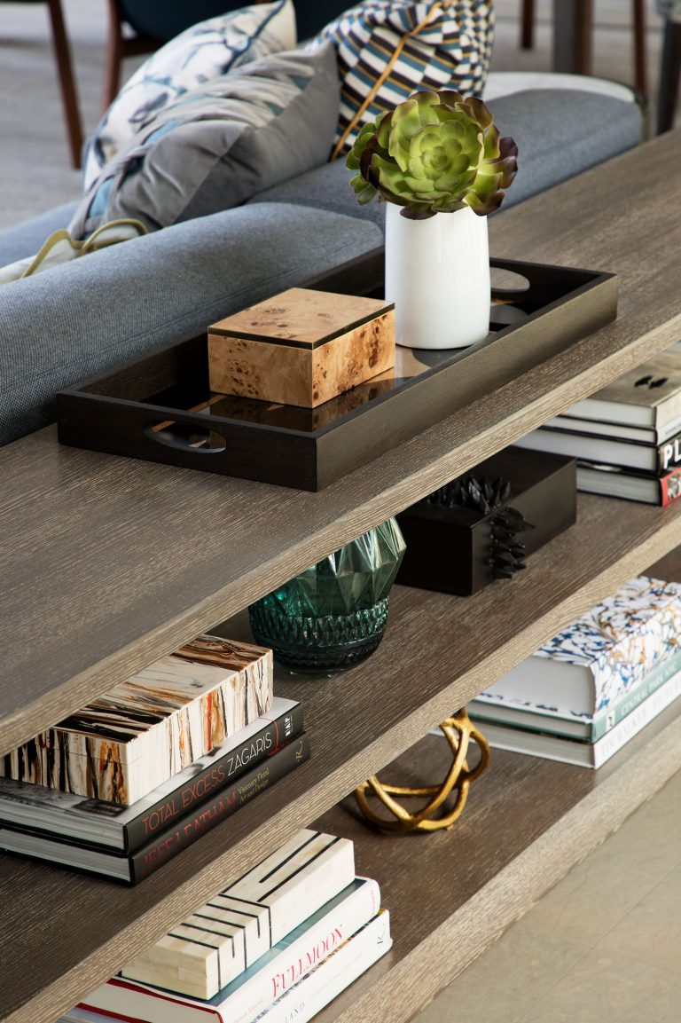A wooden shelf behind a sofa holds books, a green glass vase, decorative boxes, stacked magazines, and a black tray with a potted succulent on top. The setting appears modern and neatly arranged, with a mix of decor items and reading materials.