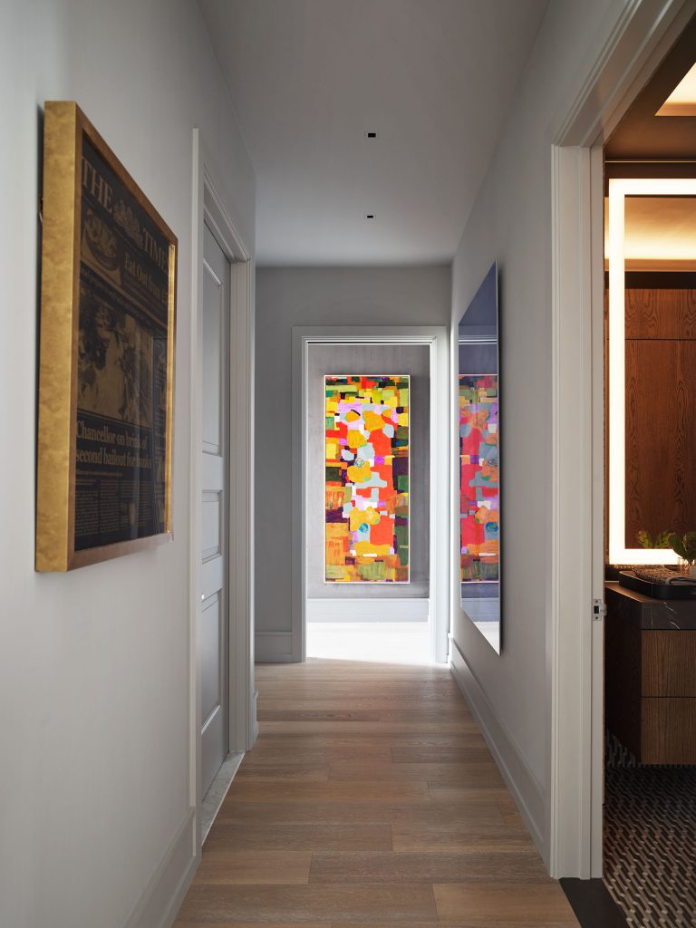 A modern hallway with light wood floors and white walls features framed art, including a colorful abstract painting at the far end. Sunlight illuminates the space, casting soft shadows. To the right, a partial view of a bathroom with warm lighting is visible.