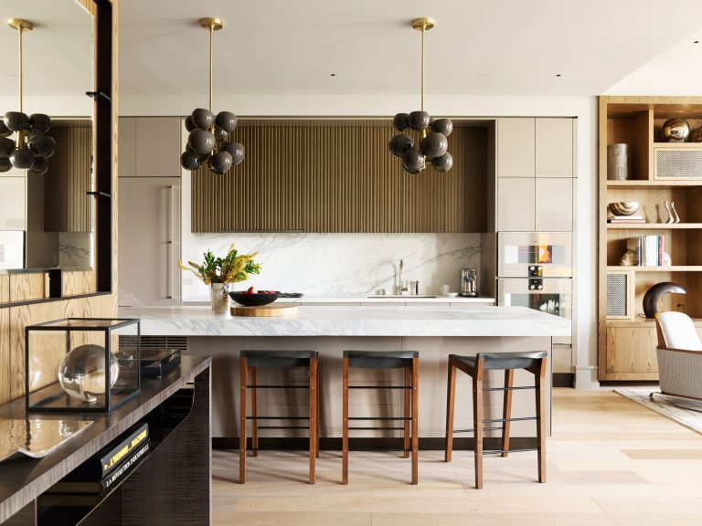 Modern kitchen with a marble island, three brown barstools, and two pendant lights with clustered black globes. Neutral cabinetry, grooved wood backsplash, and built-in appliances are visible. Open shelves with books and decor appear on the right. Flowers sit on the counter.