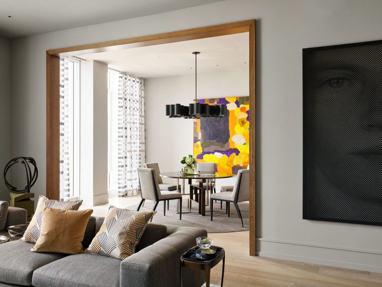 Modern living and dining space with gray sofas, patterned and tan pillows, a round dining table with six chairs, a bold abstract painting in yellow and purple, large windows with sheer curtains, and monochrome wall art featuring a partial face.