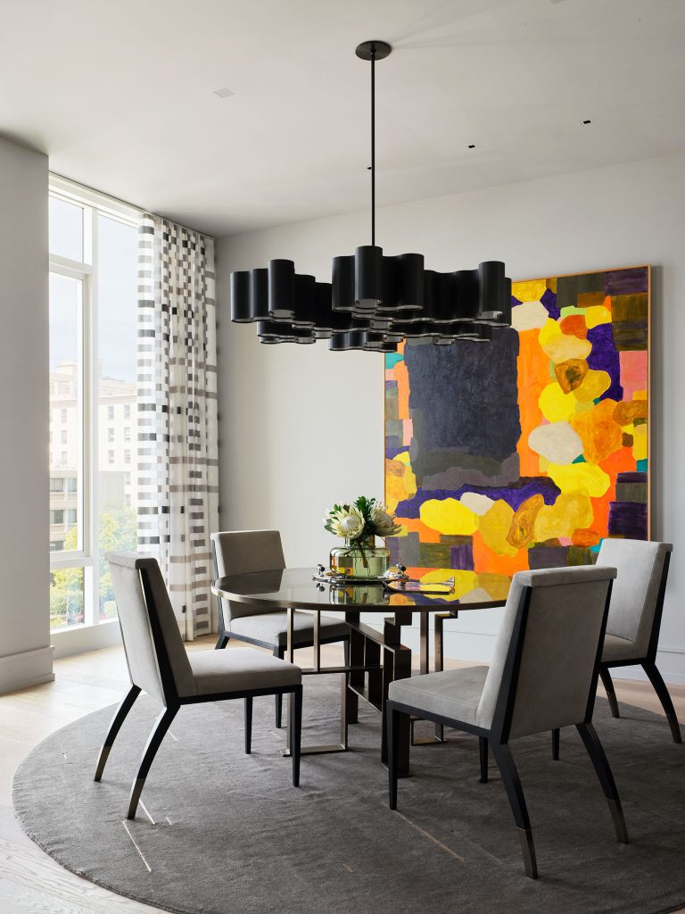 A modern dining room with four gray chairs around a circular glass table on a round gray rug. Above, a large black chandelier hangs. On the wall is a vibrant, abstract painting with yellow, orange, and purple shapes. Tall windows let in natural light.