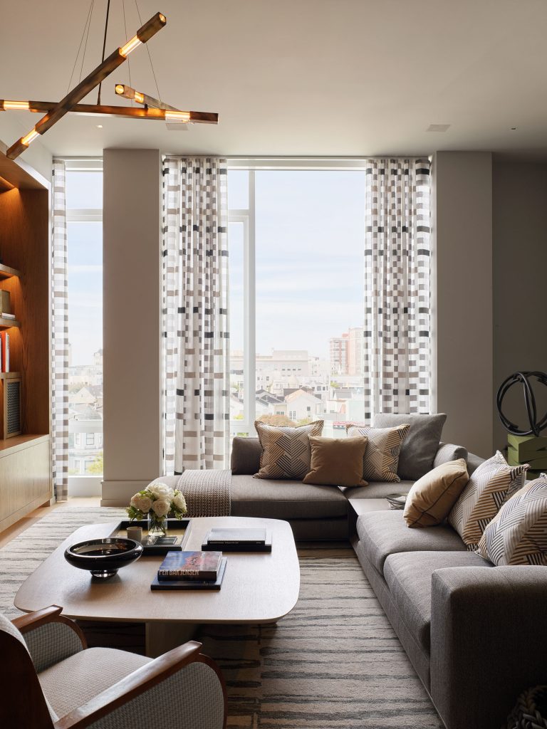 A modern living room with a large window featuring sheer, patterned curtains, a gray sectional sofa with assorted throw pillows, a wooden coffee table with books and décor, a bookshelf on the left, and a geometric chandelier overhead. Cityscape visible outside.