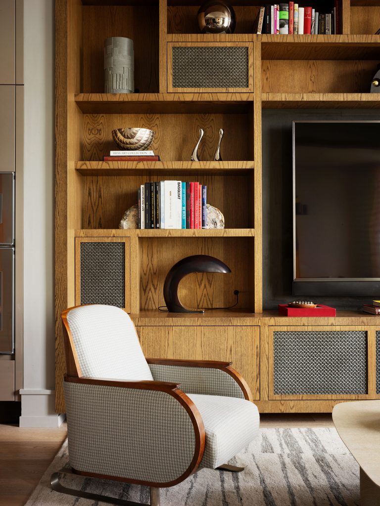 A modern living room features a checkered white armchair with wooden armrests, placed in front of a built-in wooden shelving unit. The shelves display books, decorative objects, and a TV. A patterned rug covers the floor.