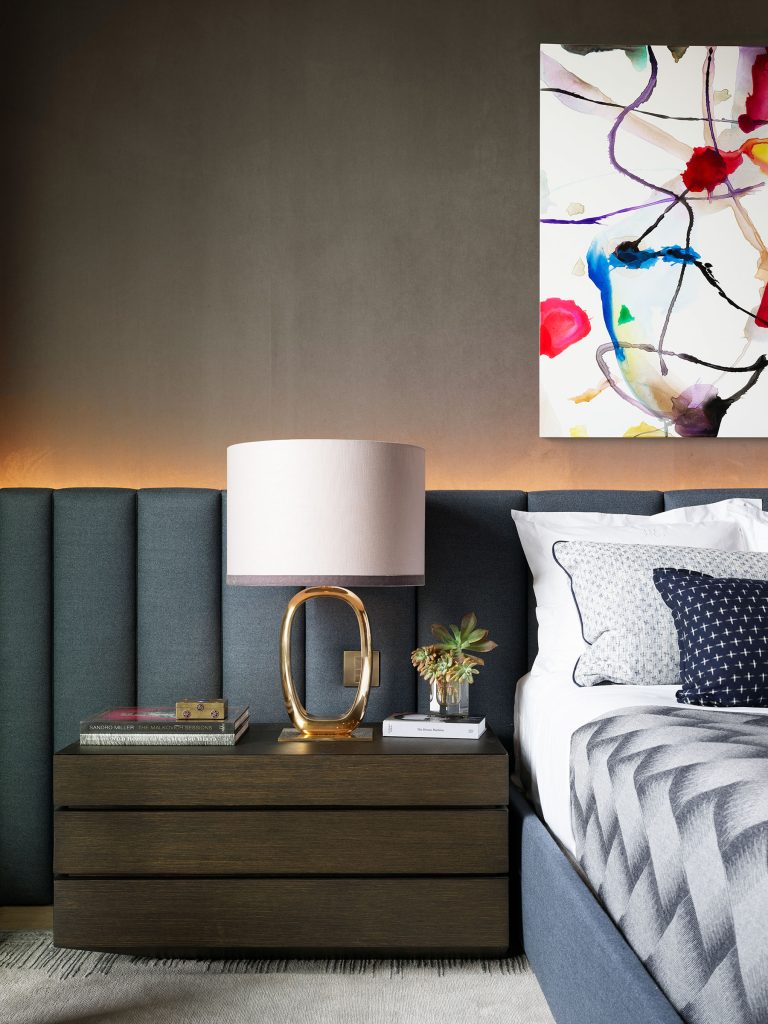 A modern bedroom scene with a grey upholstered bed, chevron-patterned bedding, and a wooden nightstand. The nightstand has a gold lamp with a white shade, stacked books, and a small plant. Above, abstract colorful artwork hangs on a dark wall with soft backlighting.
