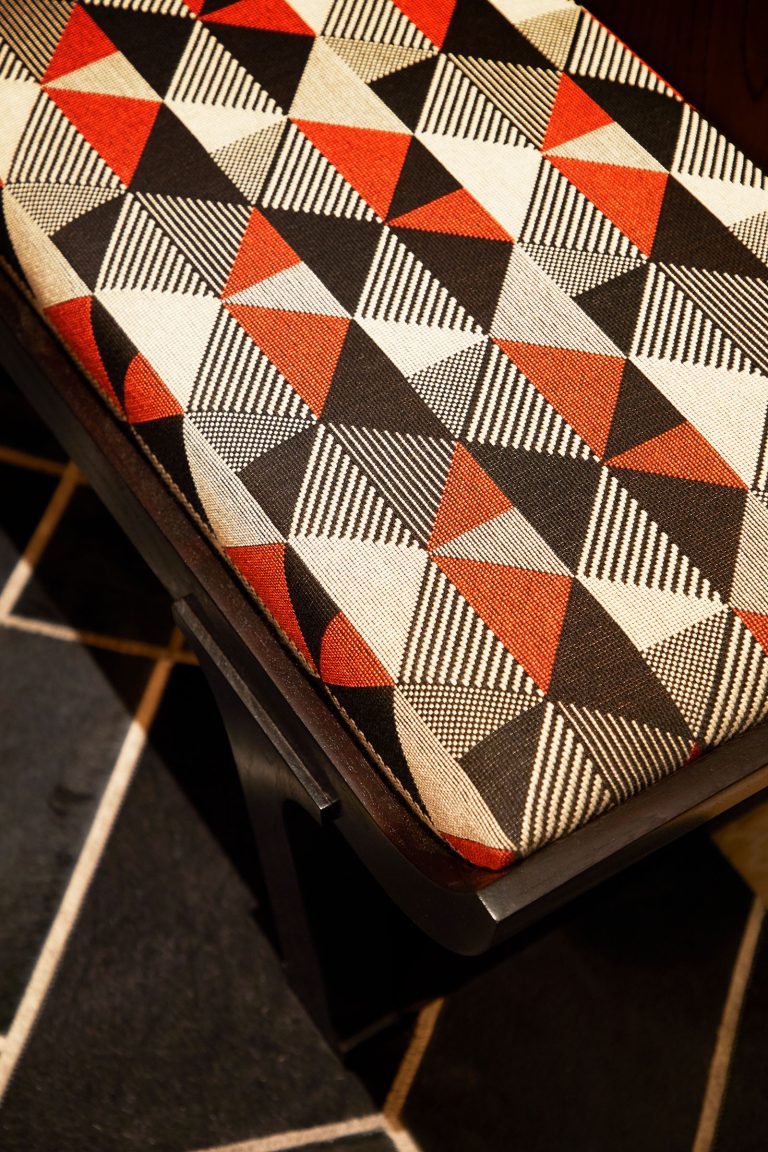 A close-up of a cushioned seat features fabric with a geometric pattern of triangles in red, black, and cream, some filled with stripes. The chair is positioned on a dark floor with a contrasting white-lined geometric design.