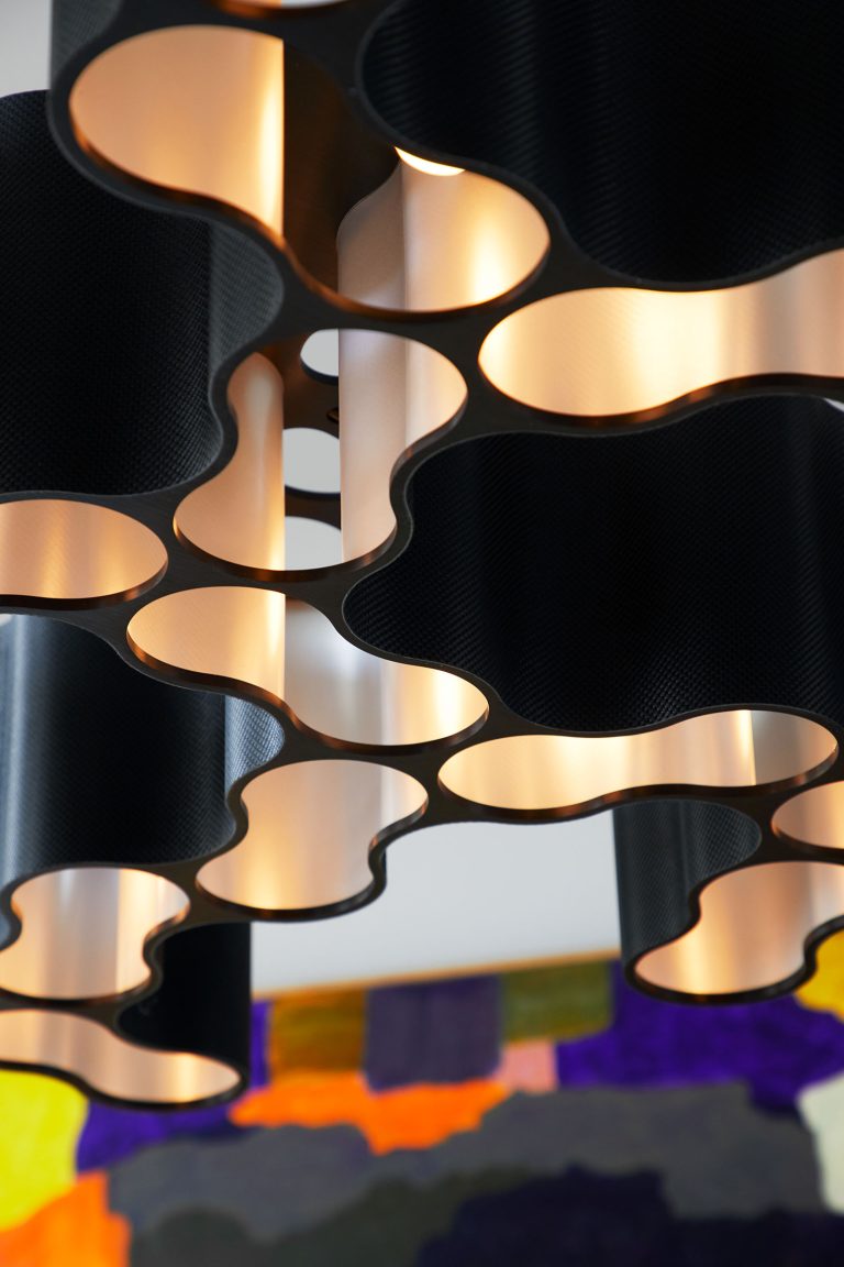 Close-up of a modern ceiling light fixture with a wavy, honeycomb-like black metal design, illuminated with warm light. In the blurred background, there is a colorful abstract painting with shades of purple, yellow, and green.