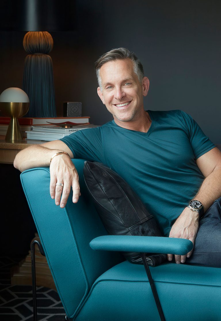 A smiling man with short gray hair and a teal shirt relaxes on a modern teal chair, leaning on a black pillow. He wears a watch and bracelet. Behind him are a lamp, books, and a dark wall, creating a cozy, stylish atmosphere.
