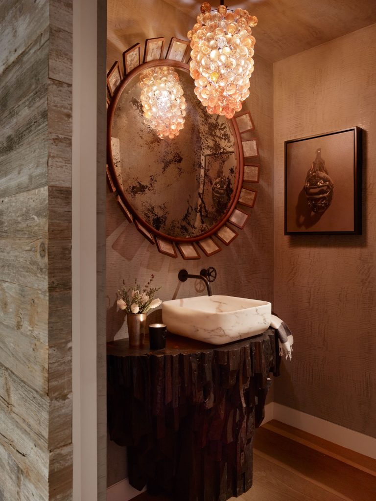 A cozy, rustic bathroom with a large round mirror framed by wood, a stone vessel sink on a dark, textured wood vanity, a glowing bubble-like glass chandelier, a small vase with flowers, and a framed art piece on the wall. Warm lighting creates a soft ambiance.