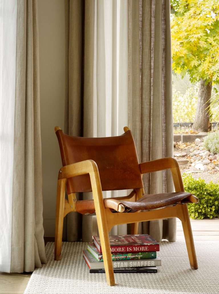 A brown leather and wood lounge chair sits on a light textured rug near floor-to-ceiling curtains. Three stacked books rest under the chair. Outside the window, green foliage and a garden are visible. Soft natural light fills the space.