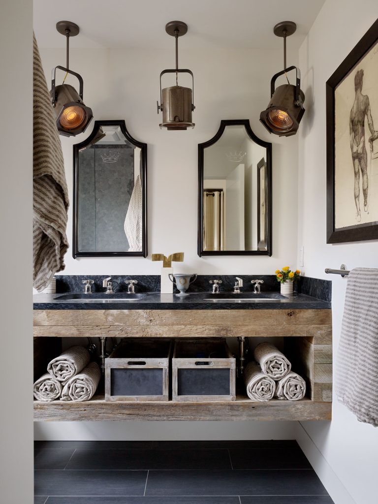 A stylish bathroom features twin mirrors with ornate black frames, a rustic wooden double sink vanity, two industrial pendant lights, rolled towels on shelves, two gray storage bins, and framed wall art of a human figure, all set against white walls.
