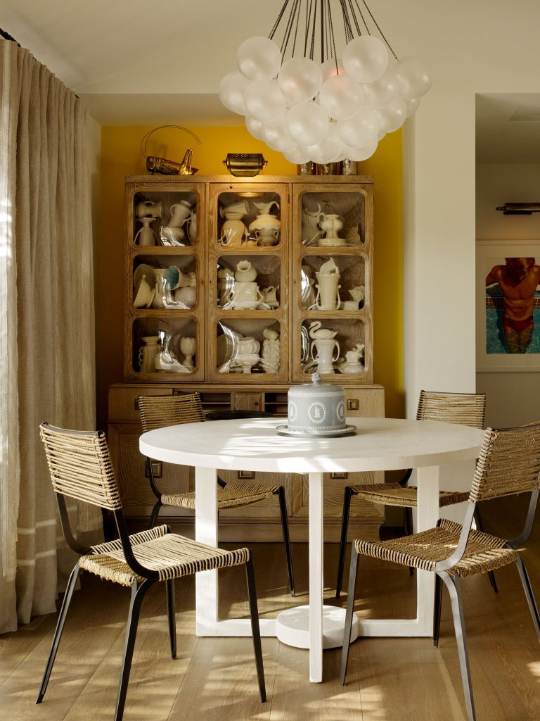 A round white table with four woven chairs sits under a modern chandelier of clustered white globes. Behind, a wooden cabinet displays various white ceramic pitchers and vessels. Light curtains and yellow wall create a bright, inviting dining area.