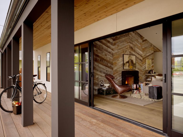 A modern porch with wooden flooring and black pillars leads to a cozy living room. The room features a chevron-patterned wooden accent wall, a brown leather chair, a fireplace, and a bicycle on the porch. A bowl of apples sits by the door.