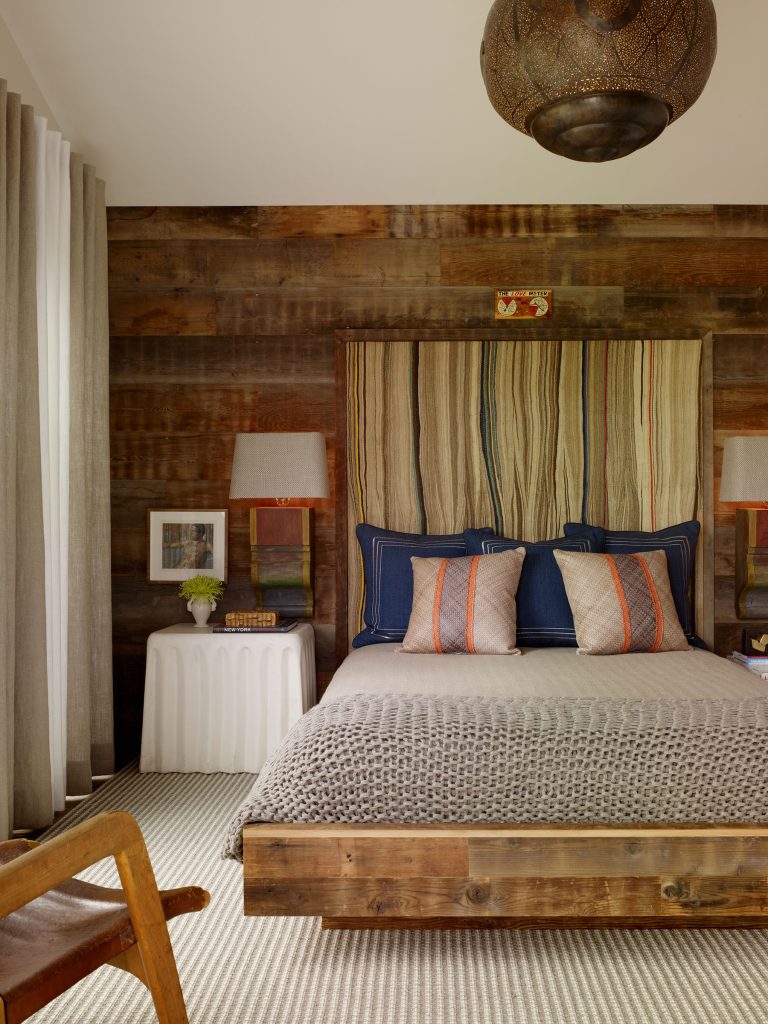 A cozy bedroom with a rustic wood accent wall, a bed with a wooden frame, blue and neutral bedding, and striped pillows. Two white side tables hold lamps and decor. Beige curtains and a woven pendant light complete the warm, inviting space.