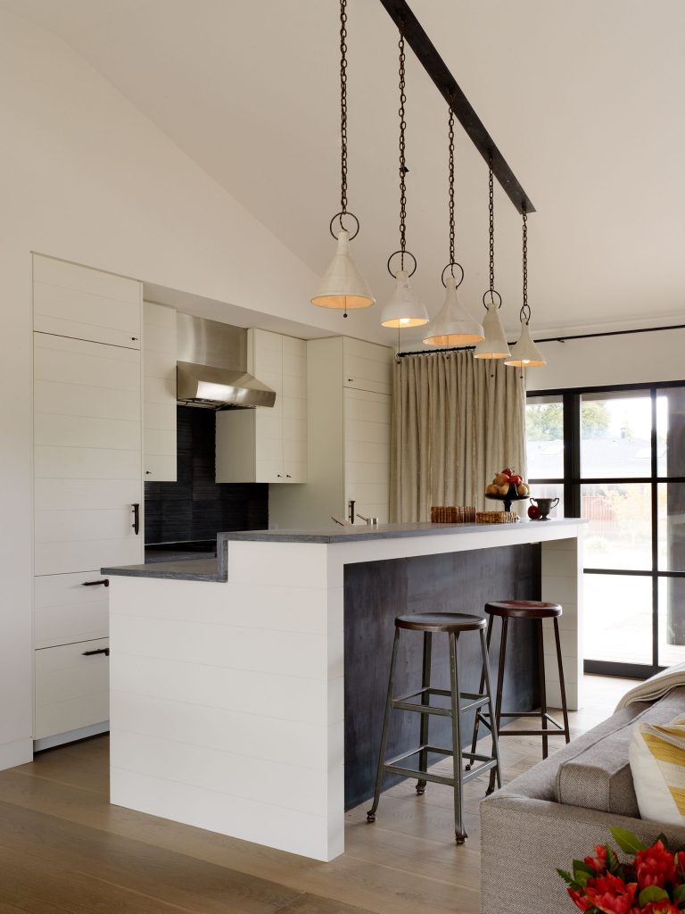 A modern kitchen with white shiplap cabinets, a black island with two metal stools, and five pendant lights hanging above. A stainless steel range hood is centered on the back wall. Light wood floors and large windows bring in natural light.