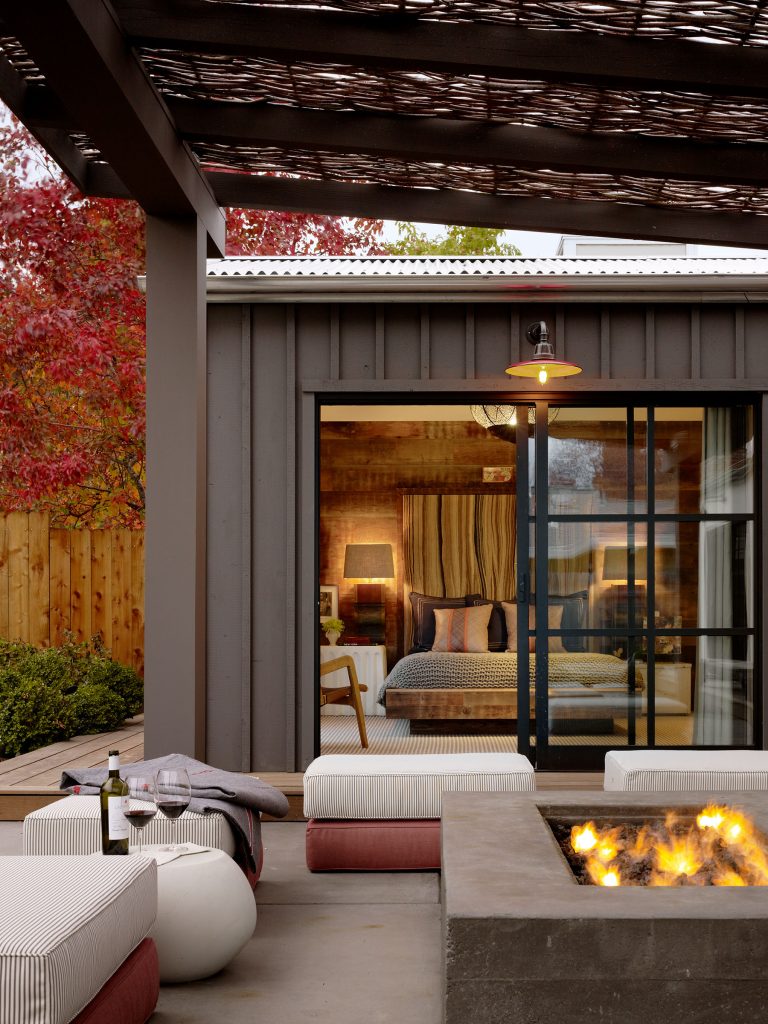 A cozy outdoor patio with cushioned seating, a concrete fire pit, a bottle of wine and glasses. Sliding glass doors reveal a warmly lit bedroom with a striped bedspread and lamps. Red autumn trees and a wooden fence are visible in the background.