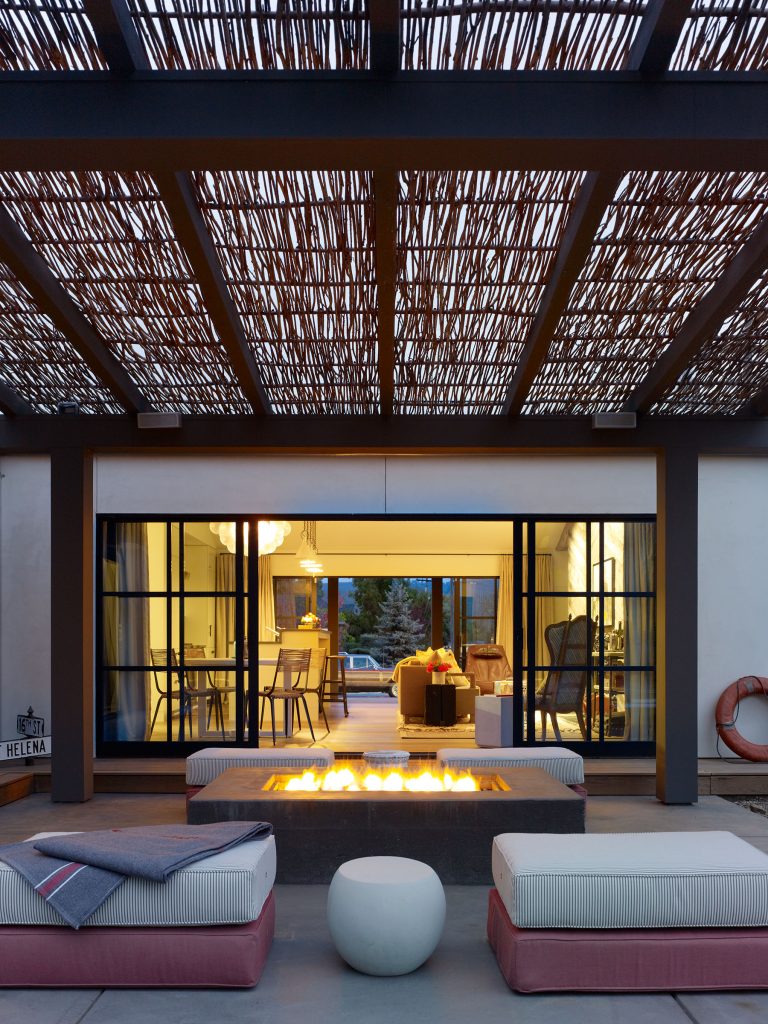 A modern outdoor patio features cushioned bench seating around a rectangular fire pit. Overhead, a woven pergola filters light. Large glass doors open to a warmly lit living space with armchairs and a couch, blending indoor and outdoor areas.