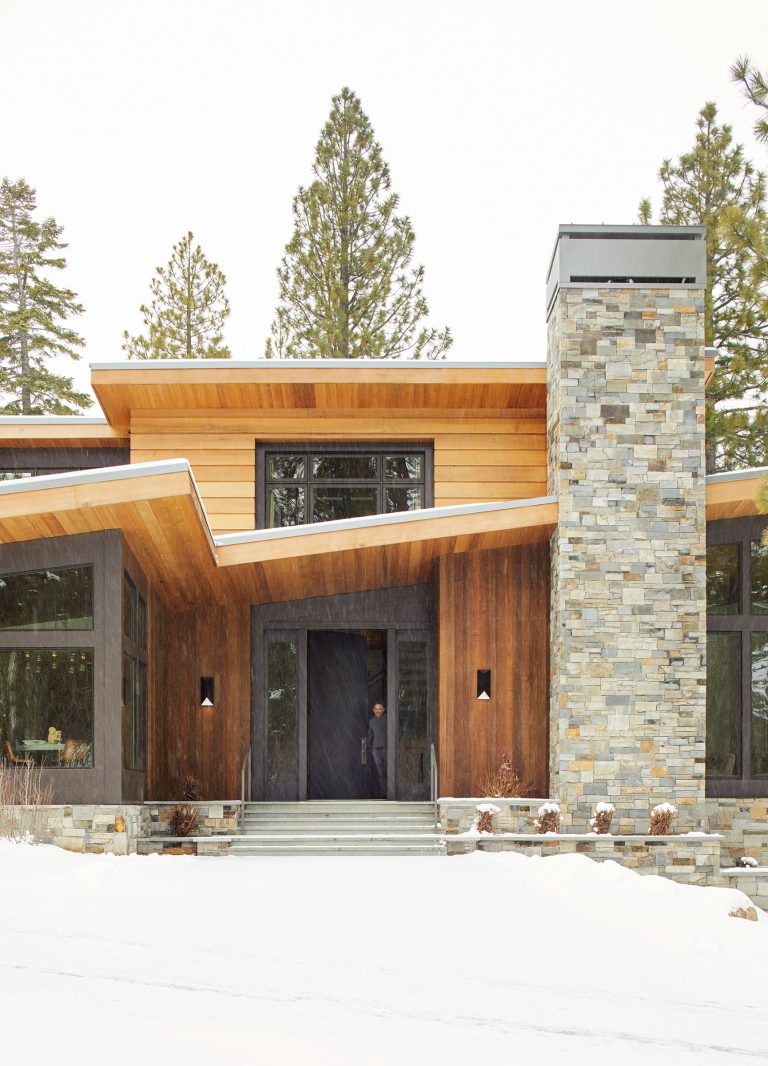 A modern house with a slanted wooden roof, large windows, and a tall stone chimney. The exterior is clad in warm wood and gray stone. Snow covers the ground and steps, and tall pine trees stand in the background.