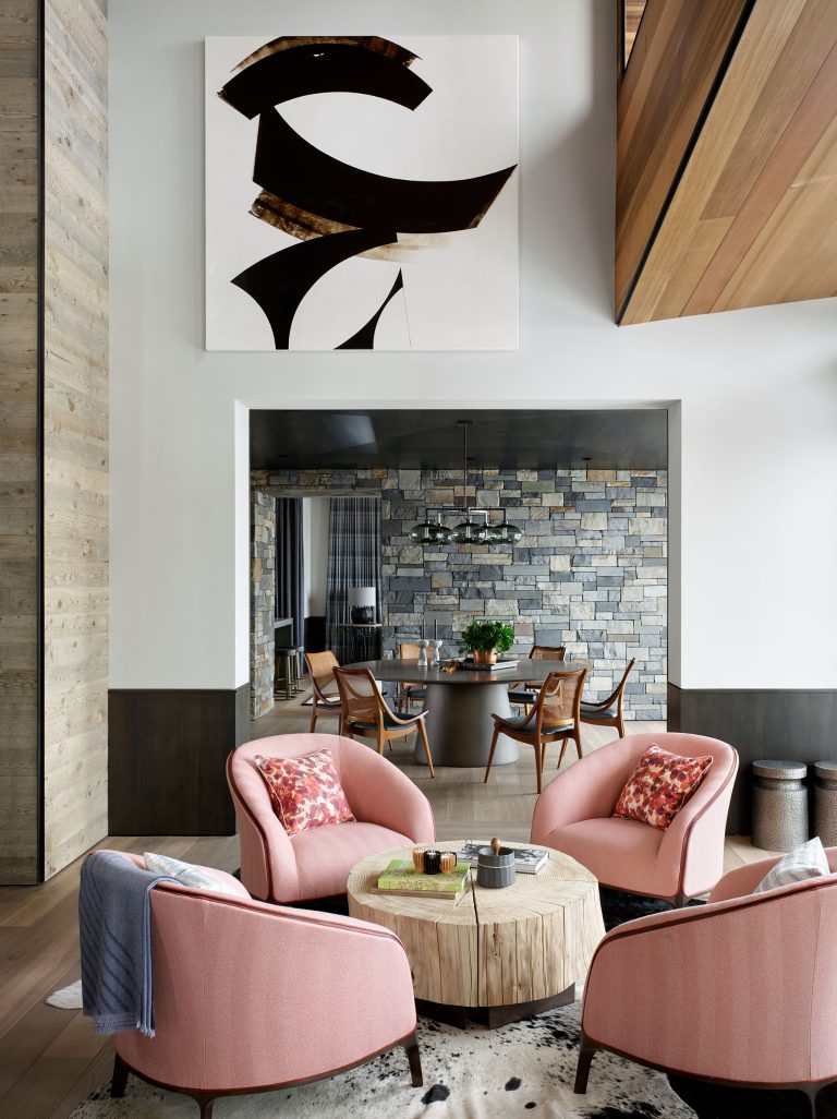 A modern living room features four pink armchairs around a round wood coffee table, set on a cowhide rug. Behind, a dining area with wood chairs and a stone wall is visible. Abstract black-and-brown art hangs above the living space. Warm, natural materials abound.
