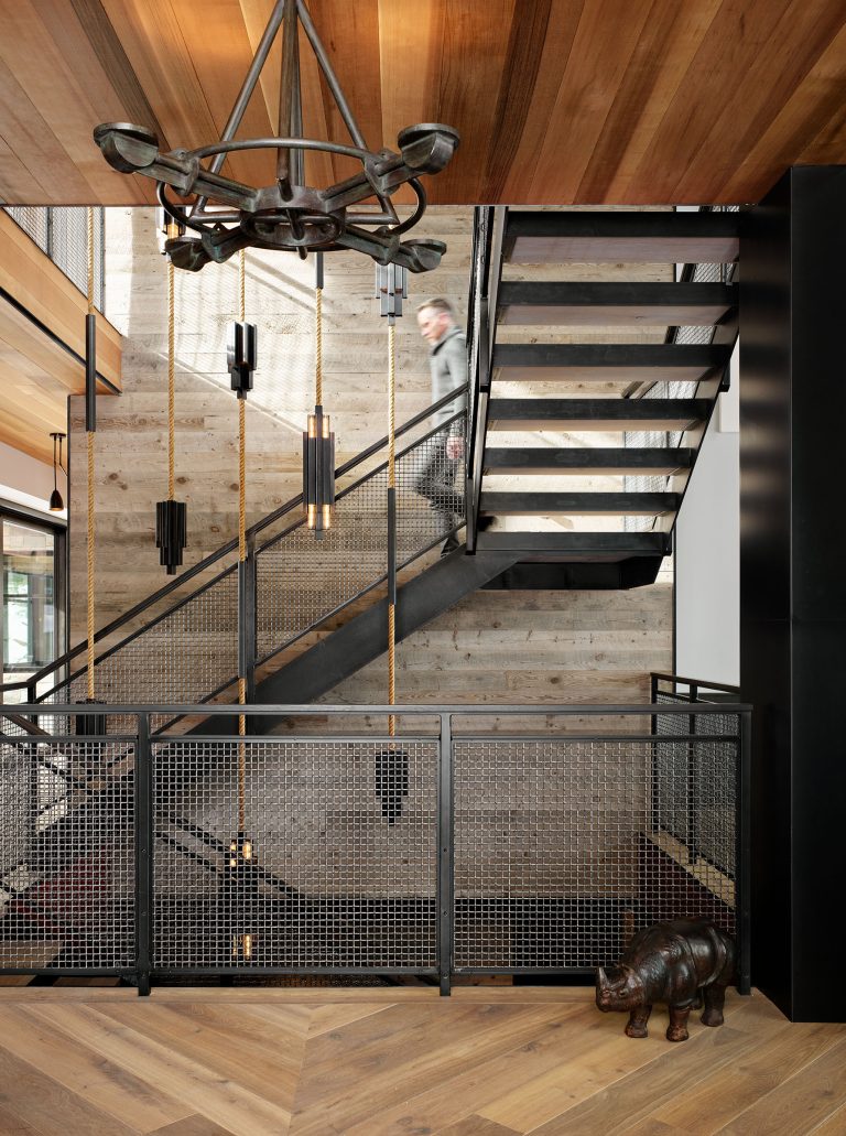 A modern interior with wood panel ceilings, a black metal chandelier, and a sleek staircase with mesh railings. Vertical pendant lights hang near the steps. A person is walking upstairs, and a decorative rhino sculpture sits by the railing on the wooden floor.