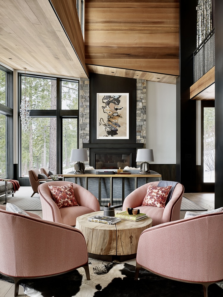 A modern living room with tall windows, light wood ceiling, and stone fireplace. Four pink armchairs with patterned pillows surround a round wood coffee table on a cowhide rug. Abstract art hangs above the mantel; two lamps sit on a console table. Tall trees visible outside.