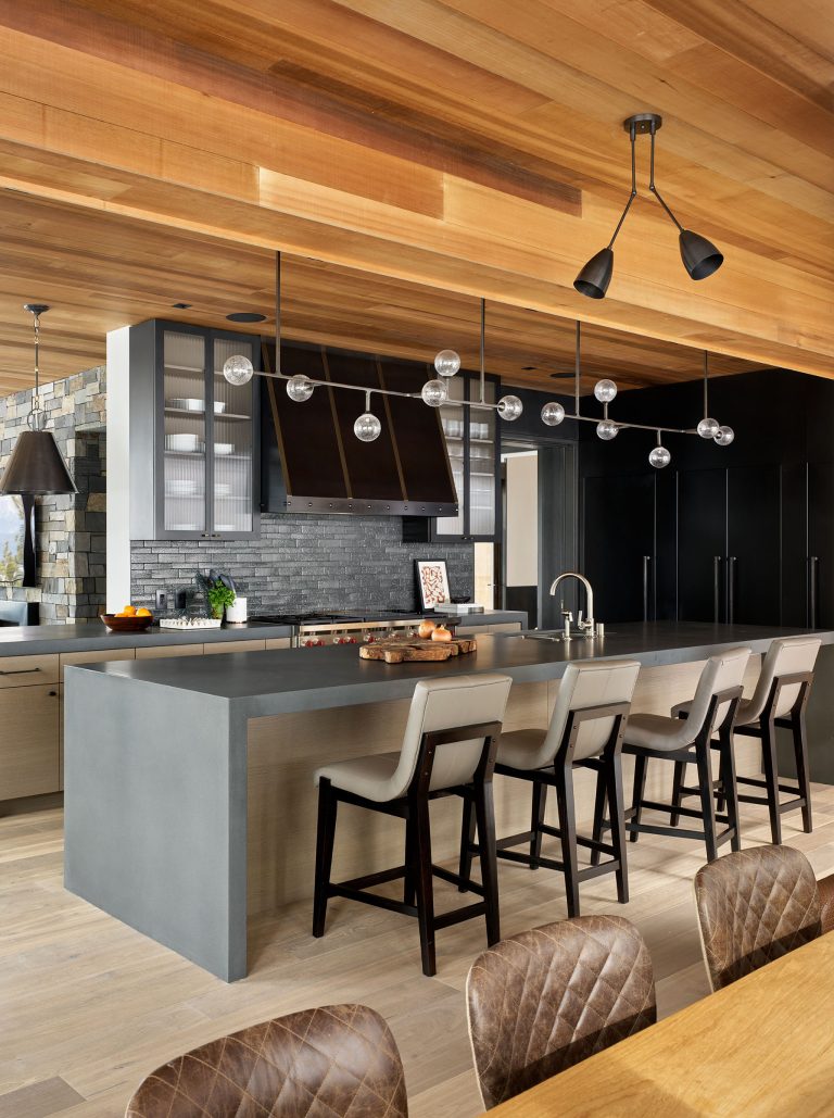 A modern kitchen with a gray island, four beige barstools, black cabinets, stainless steel appliances, and black backsplash. Globe pendant lights hang above the island. The ceiling and floor have warm wood tones, and a dining table with quilted chairs is in the foreground.