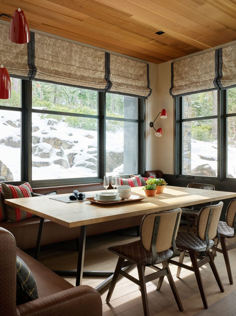 A cozy dining nook features a wooden table surrounded by padded chairs and a bench with patterned pillows. Large windows reveal a snowy landscape with rocks and trees outside. Table is set with dishes, glasses, and a small potted plant. Warm, natural wood tones prevail.