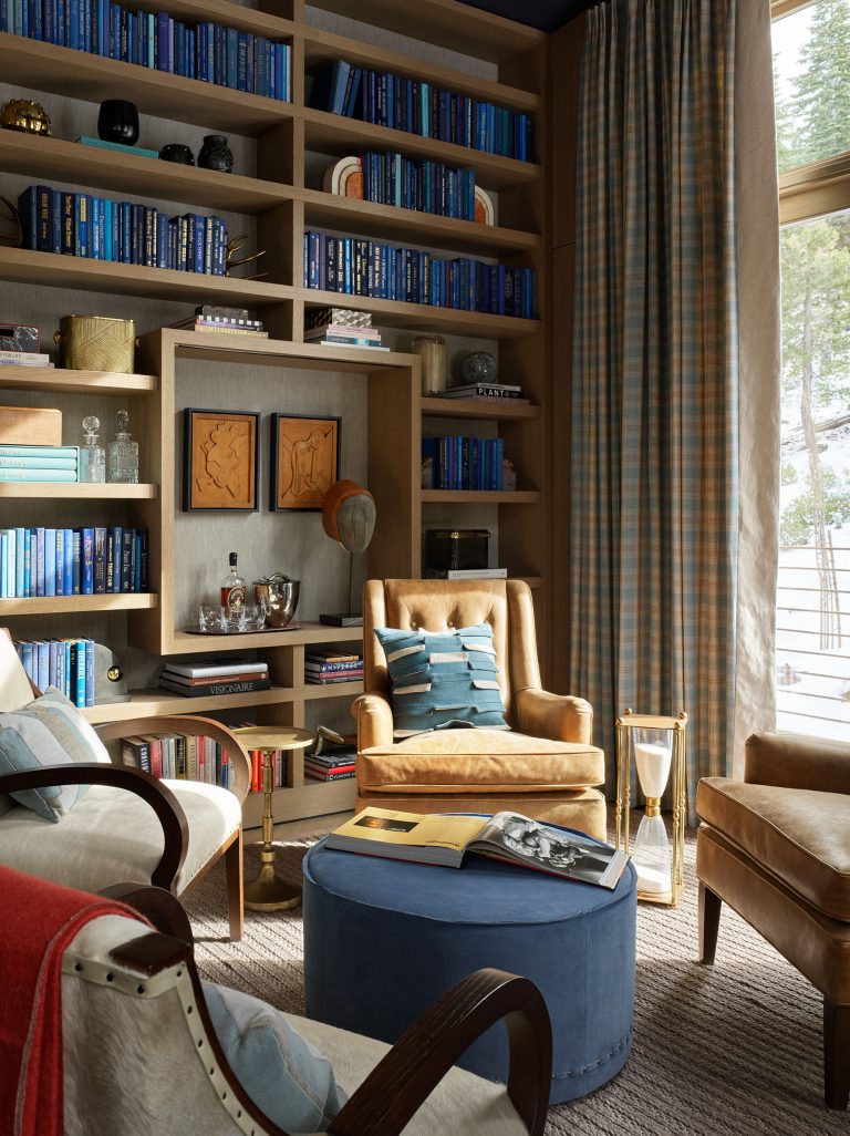 A cozy library with books organized by shades of blue on wooden shelves, tan armchairs, a round blue ottoman, a small gold side table, an hourglass, and floor-to-ceiling windows with plaid curtains overlooking a snowy outdoor scene.