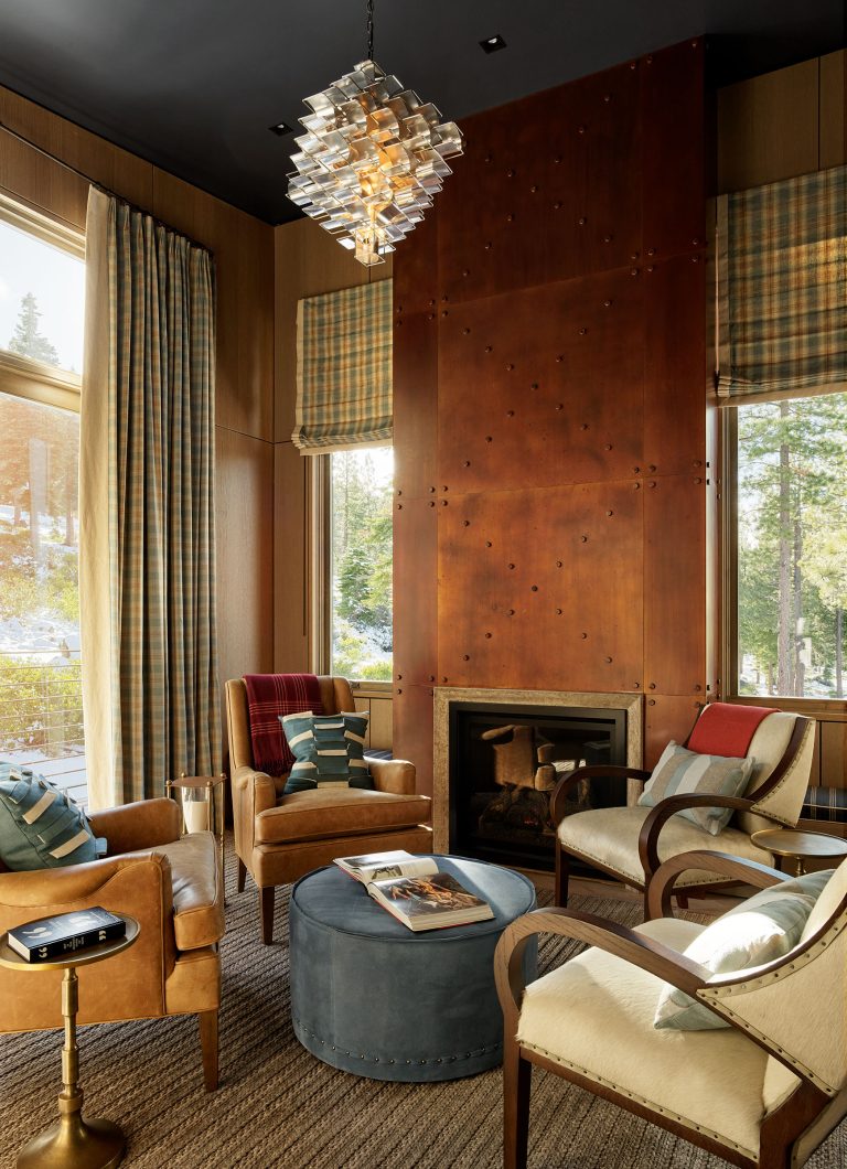 A cozy, modern living room with tan armchairs, a blue ottoman, a wooden fireplace, and large windows with plaid curtains letting in natural light. A geometric glass chandelier hangs from a dark ceiling, and trees are visible outside. Magazines rest on the ottoman.