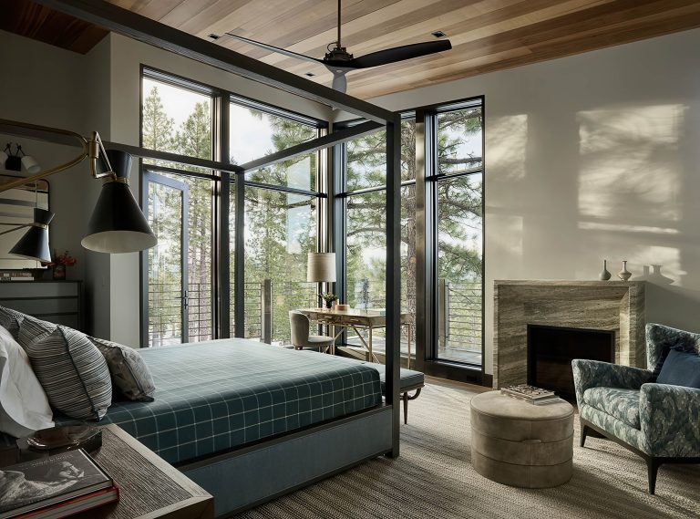 A modern bedroom with large floor-to-ceiling windows showing pine trees outside. The room has a four-poster bed with plaid bedding, a fireplace, a patterned armchair, a round ottoman, a desk near the window, and wood-accented ceiling and decor.