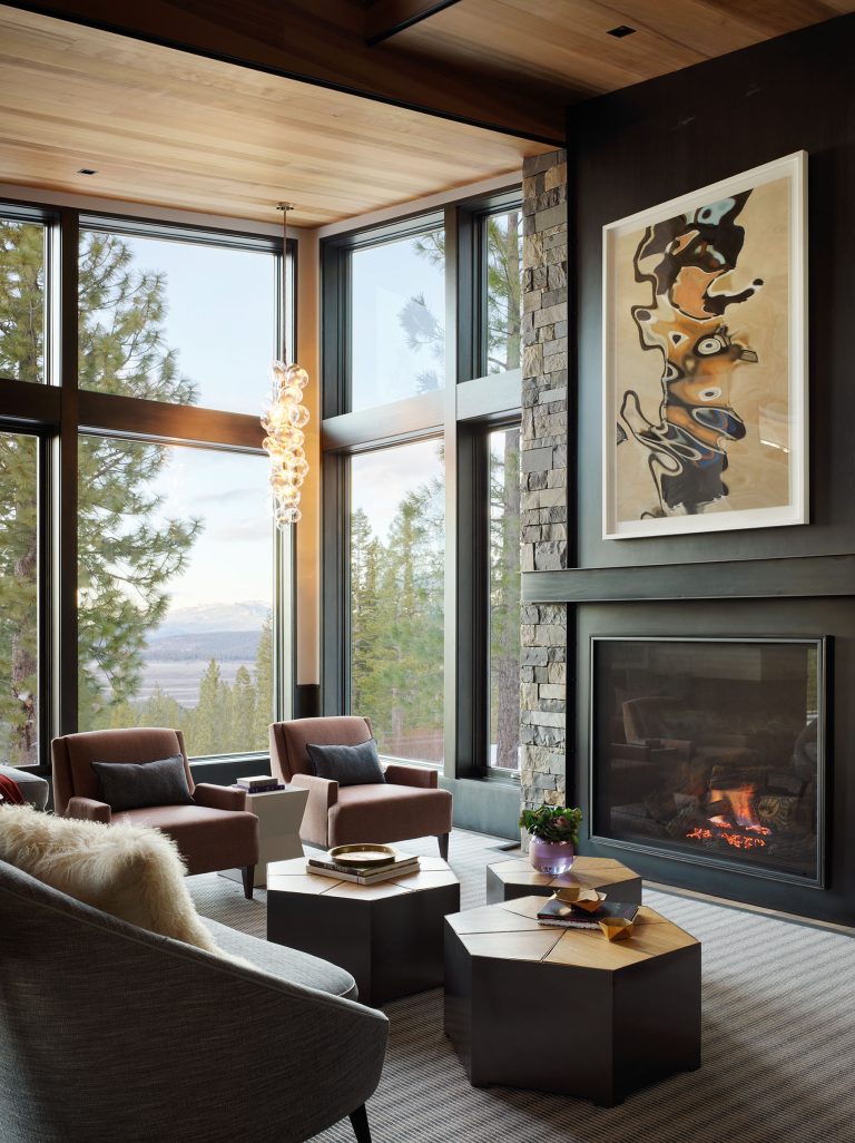 Modern living room with tall windows showing a forest view. Two brown armchairs and a furry chair face a geometric coffee table. A stone fireplace with a burning fire is topped with abstract art. The room features wood ceilings and a hanging glass light fixture.