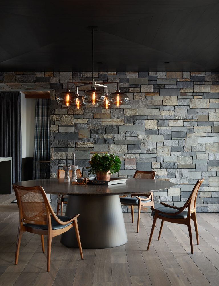 A modern dining room features a round metal table surrounded by four wooden chairs with cane backs. Above the table, a chandelier with exposed Edison bulbs hangs. The floor is light wood, and the back wall is made of irregular gray stone blocks.