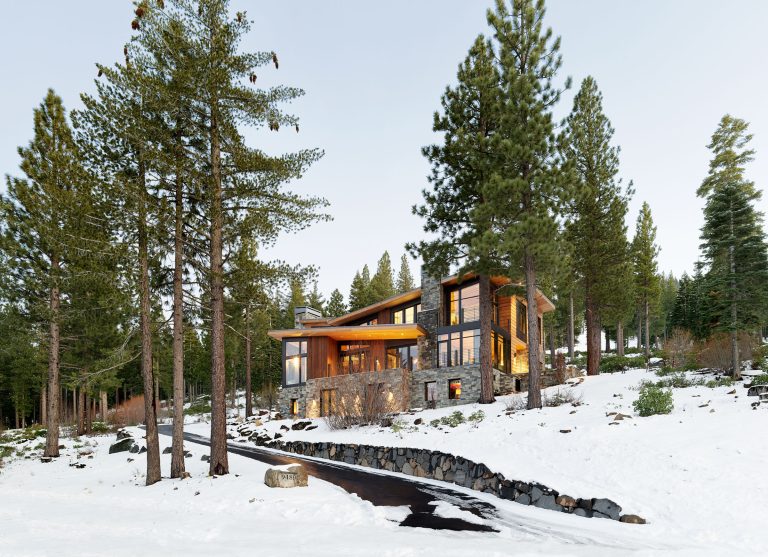 A modern house with stone and wood exterior stands among tall pine trees on a snowy landscape. Large glass windows reveal warm indoor lighting. A black, curved driveway leads to the entrance, bordered by rocks and surrounded by snow and forest.