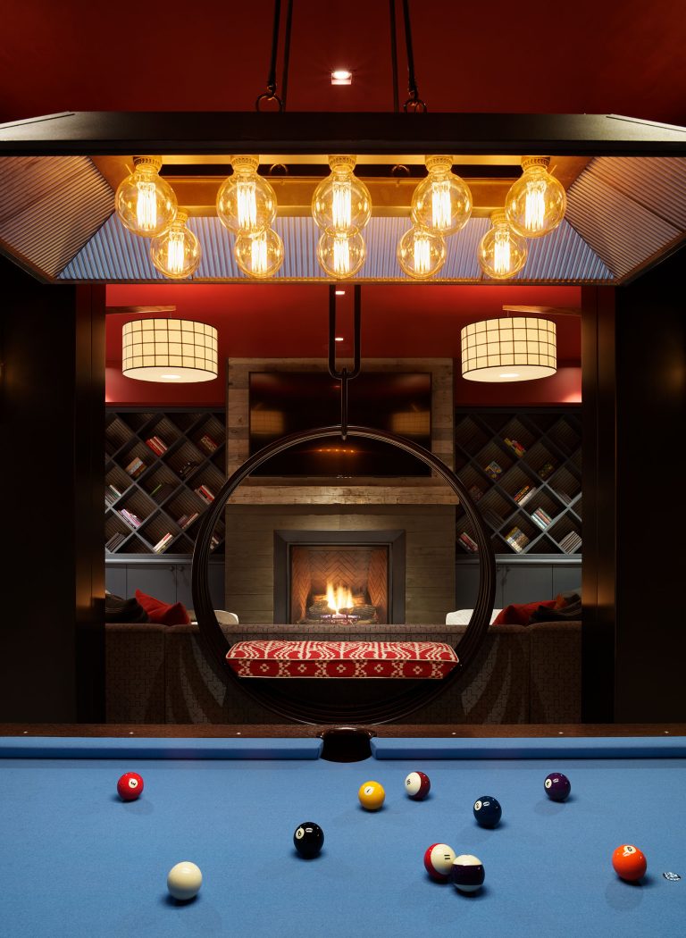 A modern room with a blue pool table in the foreground, colorful billiard balls on the table, a circular opening framing a cozy lounge with a lit fireplace, bookshelves, and stylish lighting featuring exposed bulbs above.
