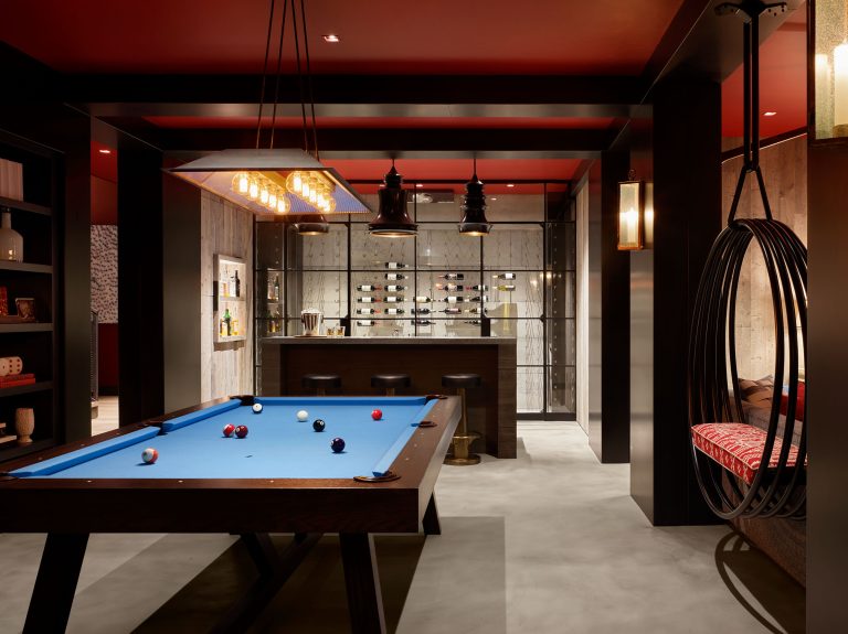 Modern game room with a blue pool table in the center, a bar with stools and wall-mounted liquor shelves at the back, hanging lights above, shelving on the left, and a hanging chair with a red cushion on the right against a concrete floor and red ceiling.
