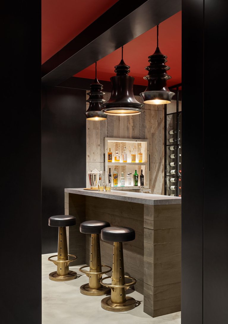 A modern home bar with a wooden counter, three brass and black barstools, hanging black pendant lights, and shelves holding liquor bottles. The ceiling is red, and the walls feature wood paneling and a stylish wine rack. The ambiance is sleek and inviting.