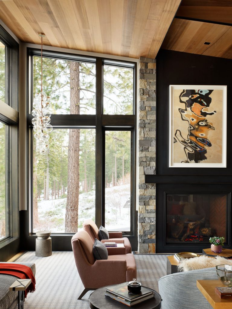 A modern living room with large floor-to-ceiling windows showing tall pine trees outside. Two pink armchairs face a stone fireplace with a black mantel. Abstract art hangs above the fire, and wooden ceilings add warmth to the space.