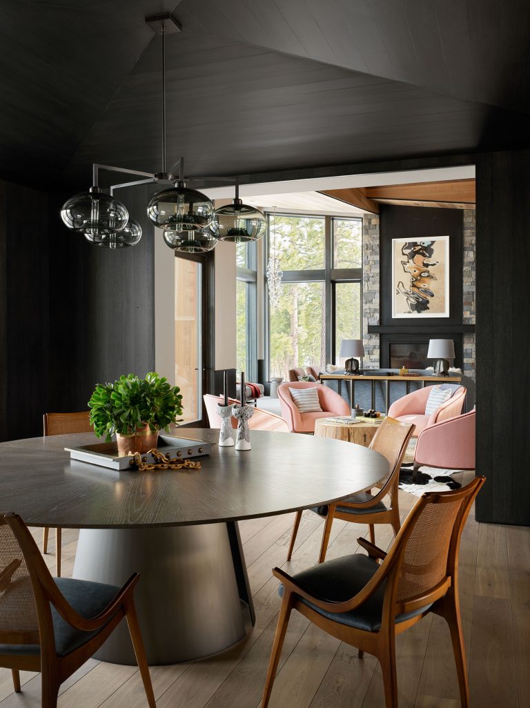 A modern dining room with a round wooden table, mid-century chairs, and hanging glass globe lights. In the background, a bright living area has pink armchairs, large windows with outdoor views, and framed art above a console table with lamps.