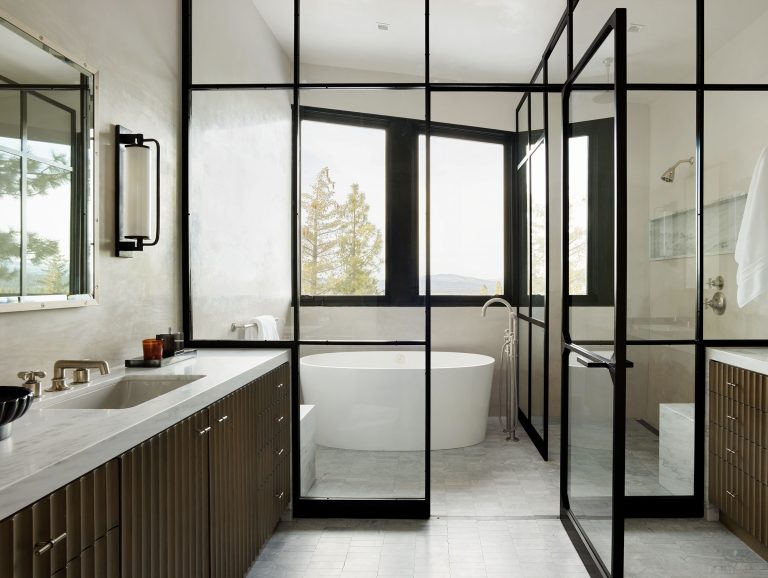 Modern bathroom with large glass panels and black frames separating the shower and bathtub areas. Freestanding white tub by a window; double sink vanity with ribbed cabinets and marble countertop; wall sconce and mirror on the left. Light, neutral color palette.