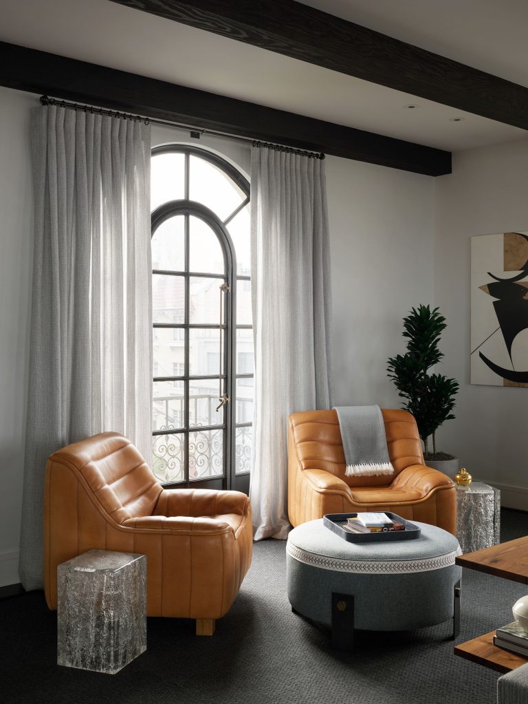A modern living room with two tan leather armchairs, a round gray ottoman, and two clear acrylic side tables. Floor-length gray curtains frame a tall arched window. A dark plant and abstract art decorate the space, which has dark flooring and exposed ceiling beams.
