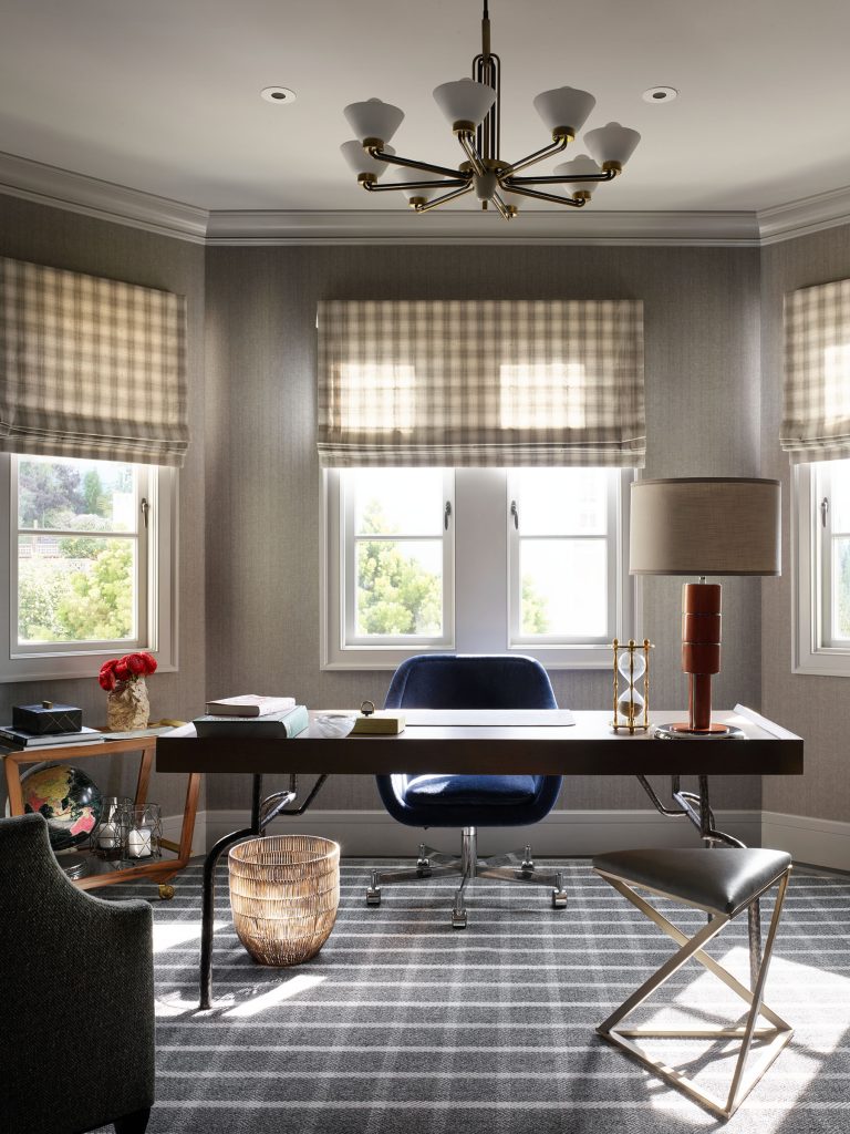 A stylish home office with a large wooden desk, navy velvet chair, geometric stool, and lamp. Three windows with plaid Roman shades let in light. A patterned rug covers the floor; a basket, red flowers, and books add cozy touches. Modern chandelier overhead.