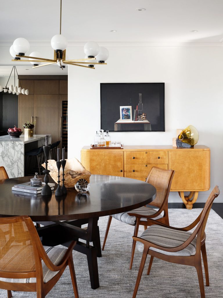 A modern dining room features a dark round table with wooden chairs, black candlesticks, and decor. Behind the table is a light wood credenza with a gold gramophone and bottles. A black-framed artwork hangs above. The room is bright with a contemporary chandelier.
