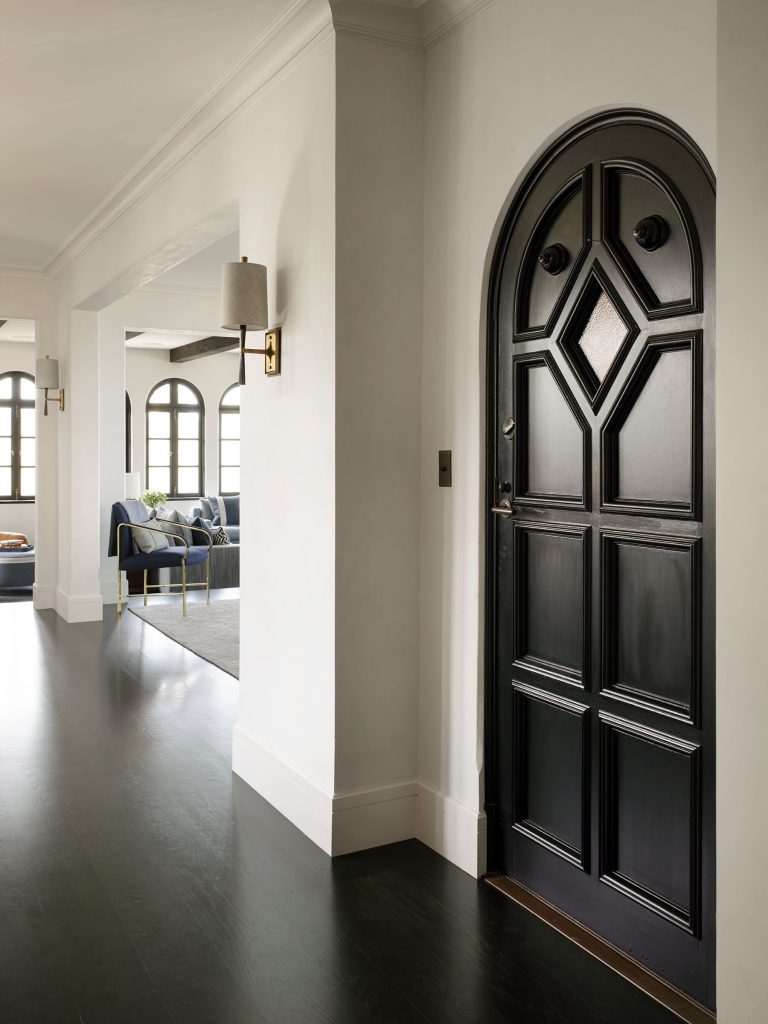 A black, arched wooden door with decorative panels and diamond-shaped window sits in a white hallway with dark wood floors. Beyond the door, a bright living space with arched windows, a blue sofa, and brass wall sconces is visible.