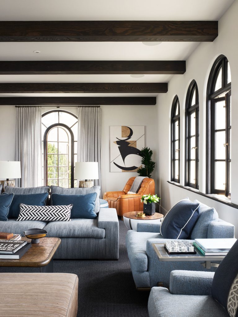 A stylish living room has arched windows with black frames, wooden ceiling beams, blue sofas, a wooden coffee table, and a caramel leather armchair in the corner. Art, plants, lamps, books, and pillows add warmth and modern decor. Light streams in from the windows.