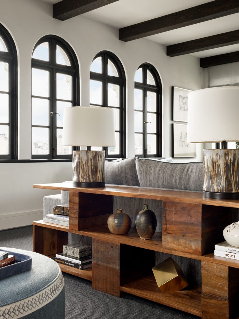 A cozy living room with arched black-framed windows, a gray sectional sofa, a wooden console table holding two decorative lamps, ceramic vases, books, and a gold sculpture. Soft natural light fills the space, highlighting modern and rustic decor.